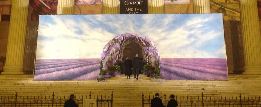 Steps of an old classic-architecutre building with large columns - with a picture of purple fields stretched across the front, with two people dressed in evening wear entering thru the middle of it…