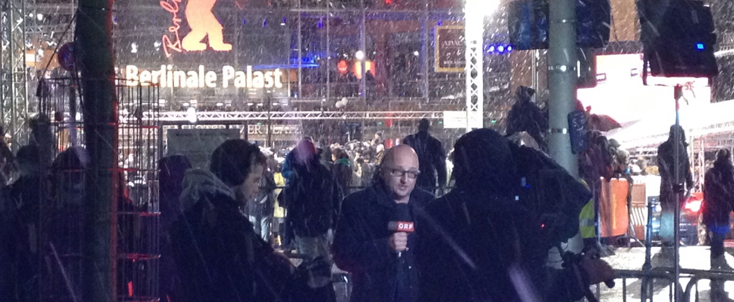 A bald headed newsreader with microphone in the snow outside of a cinema with Berlinale Palast and a red bear behind him,