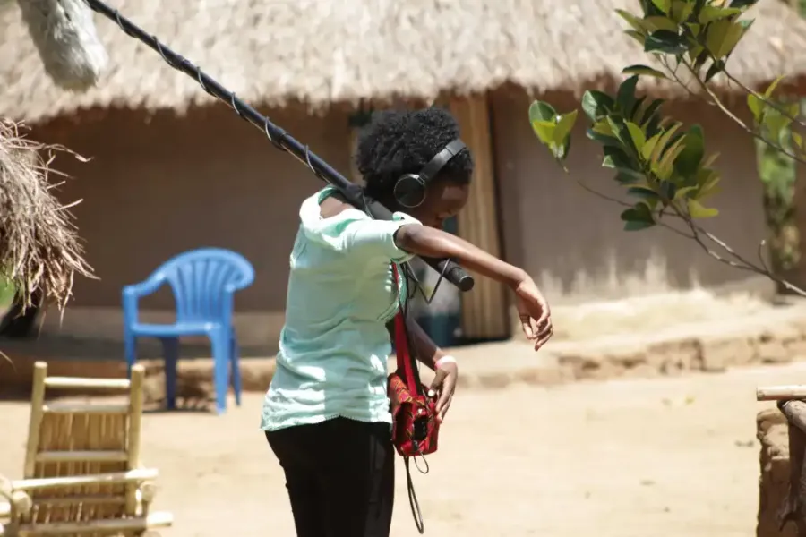 Woman boom operator, grass-roofed building in the background, sandy track.