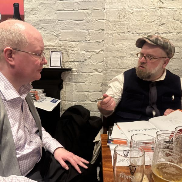 Eric Dubois signing copies of the Carnal Cinema book on a pub table with Andrew Lowes. Both wear white shirts and waist-coats.