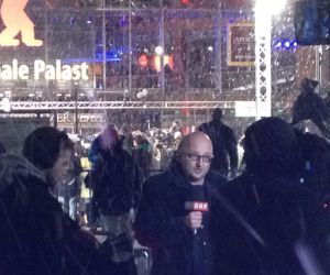 A bald headed newsreader with microphone in the snow outside of a cinema with Berlinale Palast and a red bear behind him,